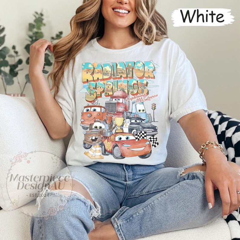 May include: White t-shirt with a graphic print of cartoon cars from the movie Cars. The text "Radiator Springs" is printed in large letters above the cars. The text "Lightning McQueen" is printed in smaller letters below the cars.