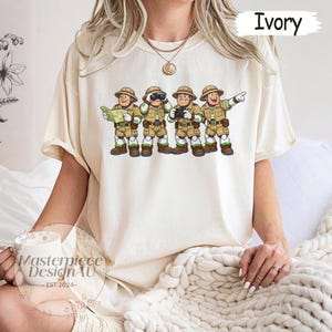 Peut inclure: T-shirt ivoire avec une illustration de dessin anim&eacute; de quatre explorateurs en tenues et chapeaux kaki. L'un tient une carte, un autre des jumelles, un troisi&egrave;me un appareil photo et le dernier pointe vers l'avant. Le texte "Ivory" est en haut &agrave; droite.