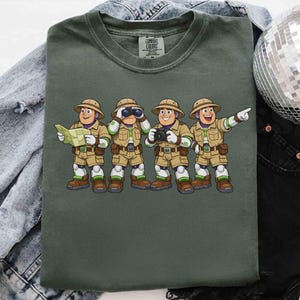 Peut inclure: T-shirt vert olive avec une illustration de quatre personnages de safari. Chaque personnage porte des v&ecirc;tements kaki et un chapeau de safari, tenant une carte, des jumelles, un appareil photo et pointant. Le t-shirt a une &eacute;tiquette Comfort Colors.