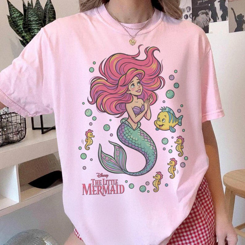 May include: A light pink crew neck t-shirt featuring a graphic print of Ariel from The Little Mermaid with long red hair, a green tail, and purple top. The design includes a yellow fish, three small yellow seahorses, and green bubbles.