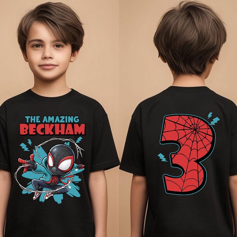 May include: Black t-shirt featuring a superhero graphic with the text "THE AMAZING BECKHAM" on the front. The back of the shirt displays a red number 3 with a spiderweb design. The shirt is worn by a child.