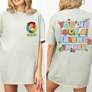 May include: A light grey t-shirt featuring a front graphic of Jessie from Toy Story inside a circle above the word NAME. The back displays a large colourful graphic with the text YOU'VE GOT A FRIEND IN ME surrounded by various Toy Story characters.