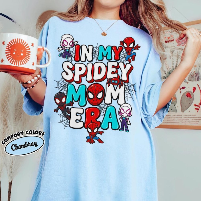 Puede incluir: Camiseta azul claro con el texto "IN MY SPIDEY MOM ERA" en letras rojas, blancas y turquesas, con personajes de Spider-Man y telara&ntilde;as. La camiseta es un Comfort Colors Chambray.