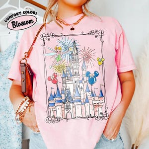 May include: Pink t-shirt with a whimsical illustration of a castle, fireworks, and Tinkerbell. The design includes Mickey Mouse balloons and a decorative border. The words "Comfort Colors" and "Blossom" are printed on the top left.