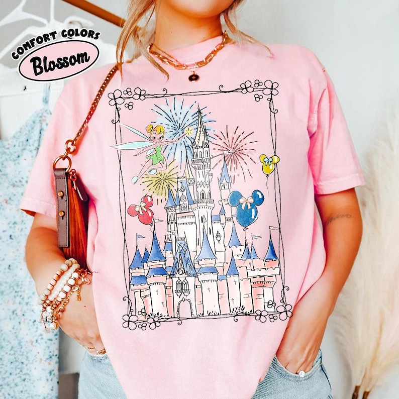 May include: Pink t-shirt with a whimsical illustration of a castle, fireworks, and Tinkerbell. The design includes Mickey Mouse balloons and a decorative border. The words "Comfort Colors" and "Blossom" are printed on the top left.