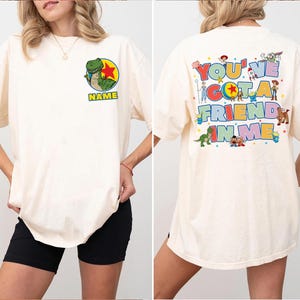 May include: An off-white t-shirt featuring a front graphic of a green dinosaur inside a yellow and red circle with the word NAME below it. The back displays various cartoon characters and the colourful text YOU'VE GOT A FRIEND IN ME.