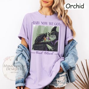 May include: Orchid-colored t-shirt featuring a graphic of a raven and a green-skinned character with the text "BABY NOW WE GOT" and "Bad Blood". The shirt is a relaxed fit and has short sleeves. The brand name "Masterpiece DesignAU" is also visible.