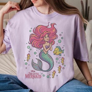 May include: A light purple t-shirt featuring a graphic print of Ariel from The Little Mermaid with long red hair, a green tail, and a purple seashell top. The design includes a yellow fish, several small yellow seahorses, and bubbles.