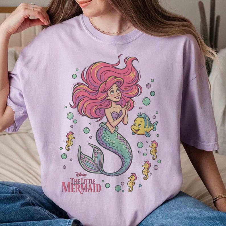 May include: A light purple t-shirt featuring a graphic print of Ariel from The Little Mermaid with long red hair, a green tail, and a purple seashell top. The design includes a yellow fish, several small yellow seahorses, and bubbles.