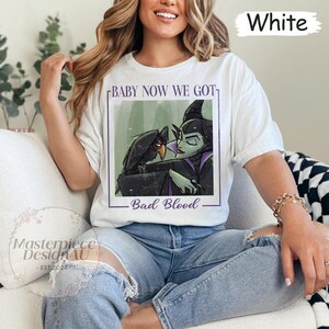 May include: White t-shirt featuring a graphic of a raven and a green-skinned character with the text "BABY NOW WE GOT - Bad Blood". The shirt is a casual style.