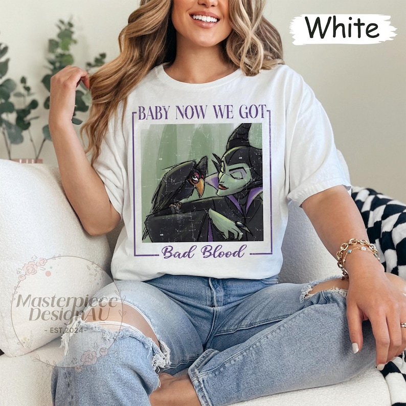 May include: White t-shirt featuring a graphic of a raven and a green-skinned character with the text "BABY NOW WE GOT - Bad Blood". The shirt is a casual style.