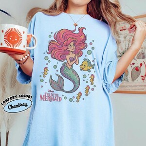 May include: A light blue t-shirt featuring a graphic of Ariel from The Little Mermaid with pink hair, a green tail, and sea creatures including Flounder and seahorses. The text reads Disney The Little Mermaid. A white mug with an orange sun design is held.