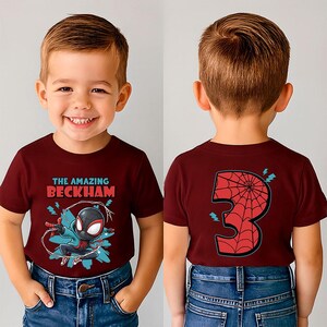 May include: Maroon t-shirt with a graphic of a superhero and the text "THE AMAZING BECKHAM" on the front. The back of the shirt features a large number "3" with a spiderweb design. The child is wearing blue jeans.