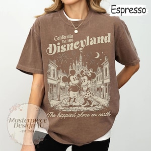 May include: A brown short-sleeve t-shirt featuring a graphic print of Mickey and Minnie Mouse in front of a castle. The text reads California Est. 1955 Disneyland and The happiest place on earth. The shirt has a crew neckline and a relaxed fit.
