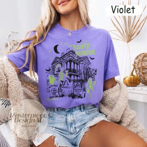 May include: Violet-colored t-shirt featuring a graphic of a haunted mansion with the text "The Haunted Mansion". The design includes bats, a moon, and tombstones with names. The shirt is a comfortable, casual wear.