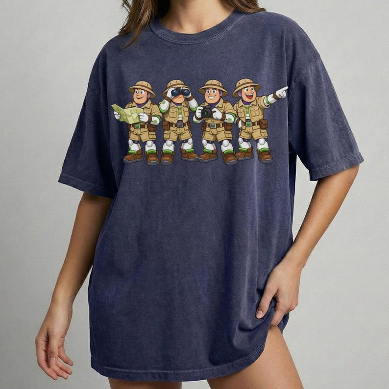 Peut inclure: T-shirt bleu marine avec une illustration de dessin anim&eacute; de quatre explorateurs. Chaque explorateur porte une tenue kaki, un chapeau et des bottes, et tient une carte, des jumelles, un appareil photo et pointe du doigt. Le t-shirt a une coupe d&eacute;contract&eacute;e.