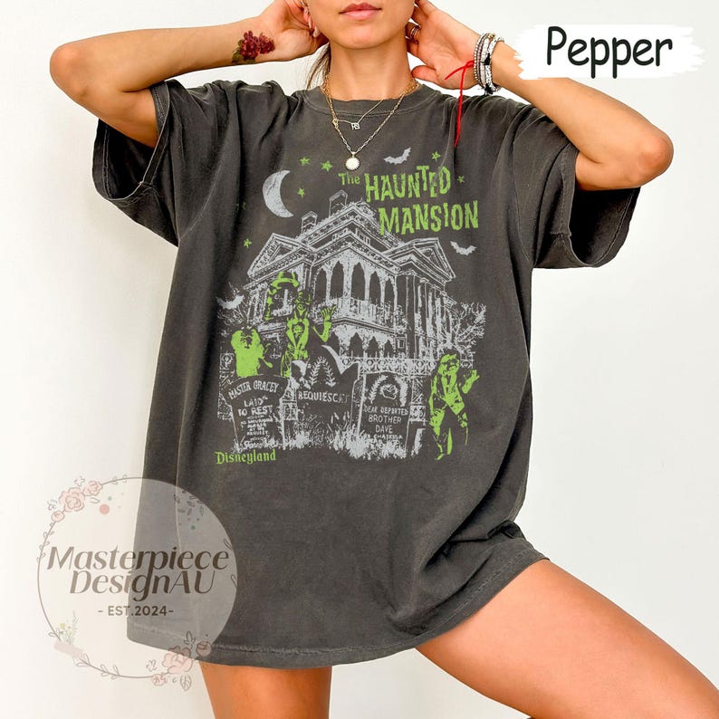 May include: Pepper-colored t-shirt featuring a graphic of The Haunted Mansion, a spooky house with green and white accents, a crescent moon, and bats. The text "The Haunted Mansion" and "Disneyland" are also visible.
