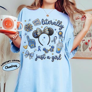 May include: Light blue t-shirt with the text "literally just a girl" and various blue and yellow graphics, including a Mickey Mouse ear headband, bows, and a foam finger. A mug with a sun design is held in the image.