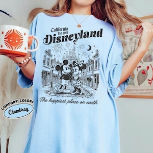 May include: A light blue t-shirt featuring a black graphic of Mickey and Minnie Mouse in front of a castle. The text reads California Est. 1955 Disneyland and The happiest place on earth. A white mug with a red sun graphic is held to the side.