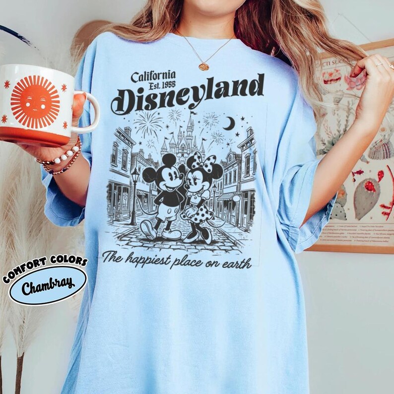 May include: A light blue t-shirt featuring a black graphic of Mickey and Minnie Mouse in front of a castle. The text reads California Est. 1955 Disneyland and The happiest place on earth. A white mug with a red sun graphic is held to the side.