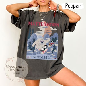 May include: Dark gray t-shirt featuring a Matterhorn Bobsleds design. The graphic includes a yeti, Mickey Mouse, and friends on skis, with the text "Matterhorn Bobsleds" in red and blue. The shirt has a vintage, worn look.