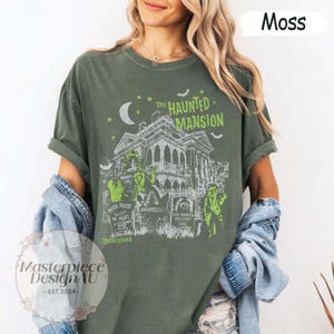 May include: Moss green t-shirt featuring a graphic of The Haunted Mansion, with a spooky house, ghosts, and tombstones. The text "The Haunted Mansion" and "Disneyland" are visible. The shirt also has the text "Masterpiece DesignAU - EST. 2024-".