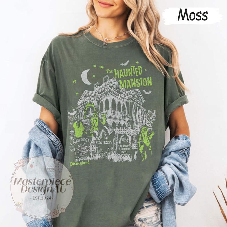 May include: Moss green t-shirt featuring a graphic of The Haunted Mansion, with a spooky house, ghosts, and tombstones. The text "The Haunted Mansion" and "Disneyland" are visible. The shirt also has the text "Masterpiece DesignAU - EST. 2024-".