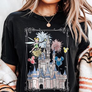 May include: Black t-shirt featuring a colorful graphic of a fairy, fireworks, and a castle. The design includes pink, blue, yellow, and white elements, with Mickey Mouse-shaped balloons. The shirt has a distressed look.