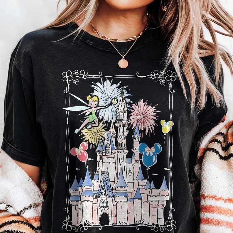 May include: Black t-shirt featuring a colorful graphic of a fairy, fireworks, and a castle. The design includes pink, blue, yellow, and white elements, with Mickey Mouse-shaped balloons. The shirt has a distressed look.