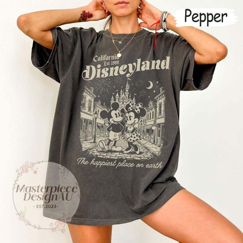 May include: A charcoal grey short-sleeve t-shirt featuring a graphic print of Mickey and Minnie Mouse in front of a castle. The text reads California Est. 1955 Disneyland and The happiest place on earth. The shirt has a crew neckline and loose fit.
