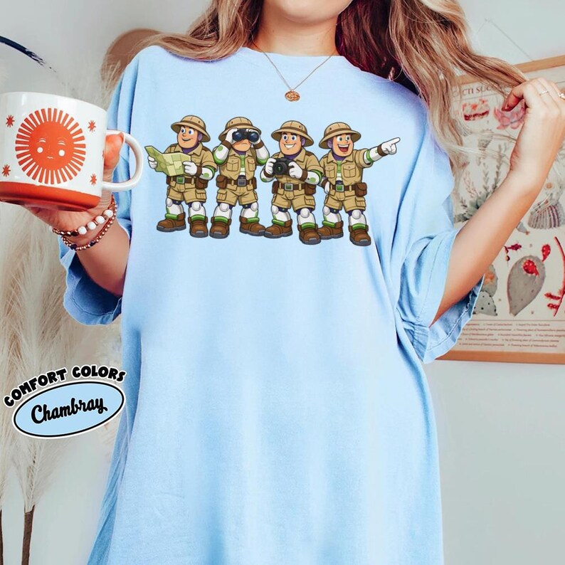 Peut inclure: T-shirt bleu clair avec une illustration de quatre explorateurs en tenues kaki, chacun avec des accessoires diff&eacute;rents. Le t-shirt porte les mots "Comfort Colors Chambray" dans un design circulaire. La personne tient une tasse avec un soleil.