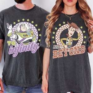 May include: Two dark gray t-shirts featuring characters from Toy Story. One shirt has Buzz Lightyear with the text "To Infinity" and the other has Woody with the text "And Beyond". Both designs are surrounded by stars and a checkered circle.