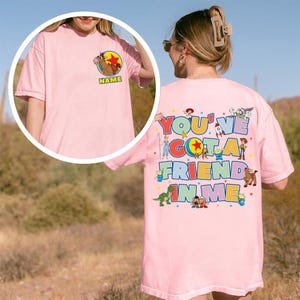 May include: A light pink t-shirt featuring a small graphic of a horse character and the word NAME on the front left chest. The back displays the text YOU'VE GOT A FRIEND IN ME surrounded by colourful illustrations of various animated toy characters.