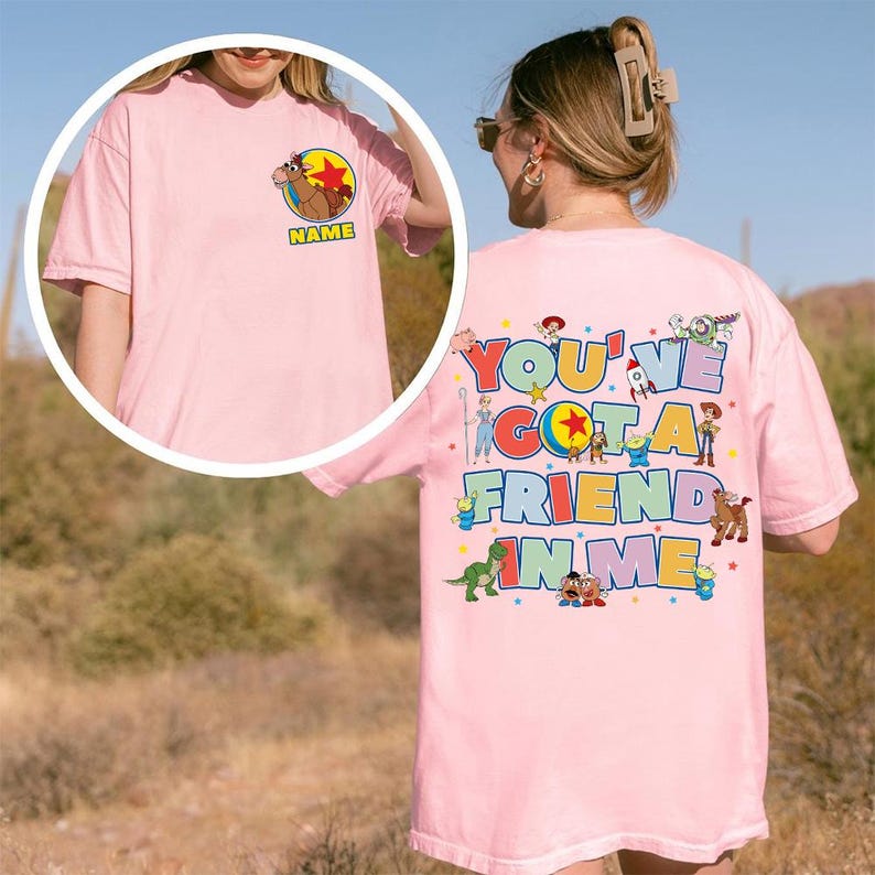 May include: A light pink t-shirt featuring a small graphic of a horse character and the word NAME on the front left chest. The back displays the text YOU'VE GOT A FRIEND IN ME surrounded by colourful illustrations of various animated toy characters.