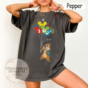 May include: Dark gray t-shirt featuring a cartoon chipmunk holding balloons that read "Happy 10th Day of School". The balloons are red, yellow, green, and blue. The shirt has the text "Name" and the logo "Masterpiece DesignAU - EST.2024-".