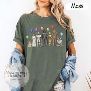 May include: Moss green t-shirt with a Star Wars character design. The graphic shows characters such as Darth Vader and R2-D2, each with a balloon. The shirt also has the text "Masterpiece Design AU - EST. 2024".