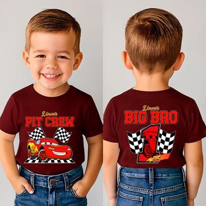 May include: A maroon t-shirt featuring a cartoon race car with the text "Liam's PIT CREW" and checkered flags. The back of the shirt says "Liam's BIG BRO" with a race car and checkered flags. The shirt is worn by a young child.