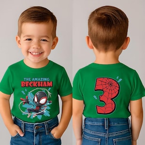 May include: A green t-shirt featuring a cartoon superhero graphic with the text "THE AMAZING BECKHAM" in red and white. The back of the shirt displays a red number "3" with a spiderweb design. The child is wearing blue jeans.