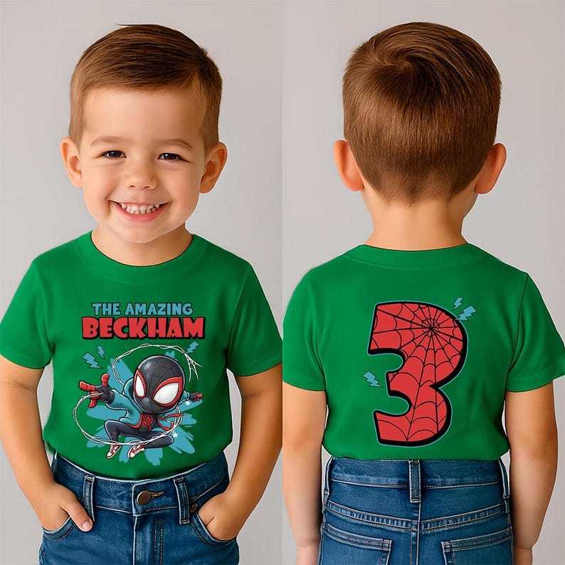 May include: A green t-shirt featuring a cartoon superhero graphic with the text "THE AMAZING BECKHAM" in red and white. The back of the shirt displays a red number "3" with a spiderweb design. The child is wearing blue jeans.
