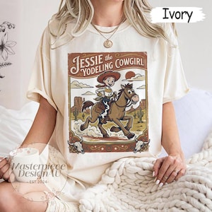 May include: An ivory short-sleeve t-shirt featuring a graphic print of a cowgirl riding a horse in a desert landscape. The text reads Jessie the Yodeling Cowgirl. The design includes cacti, rock formations, and two steer skulls at the bottom corners.