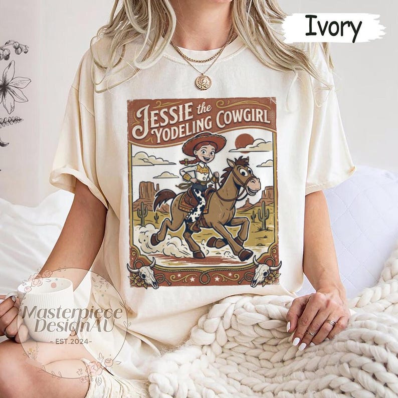May include: An ivory short-sleeve t-shirt featuring a graphic print of a cowgirl riding a horse in a desert landscape. The text reads Jessie the Yodeling Cowgirl. The design includes cacti, rock formations, and two steer skulls at the bottom corners.