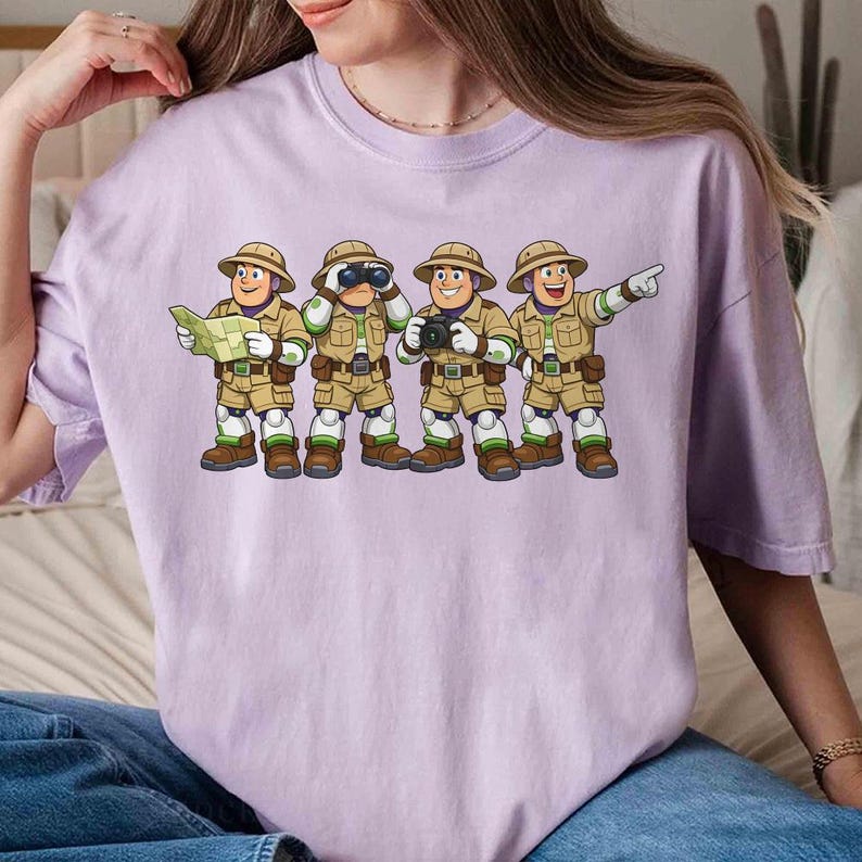 Peut inclure: T-shirt lilas clair avec une illustration de quatre explorateurs en tenues et chapeaux kaki. L'un tient une carte, un autre des jumelles, un troisi&egrave;me un appareil photo et le dernier pointe vers l'avant. Les explorateurs sont align&eacute;s.