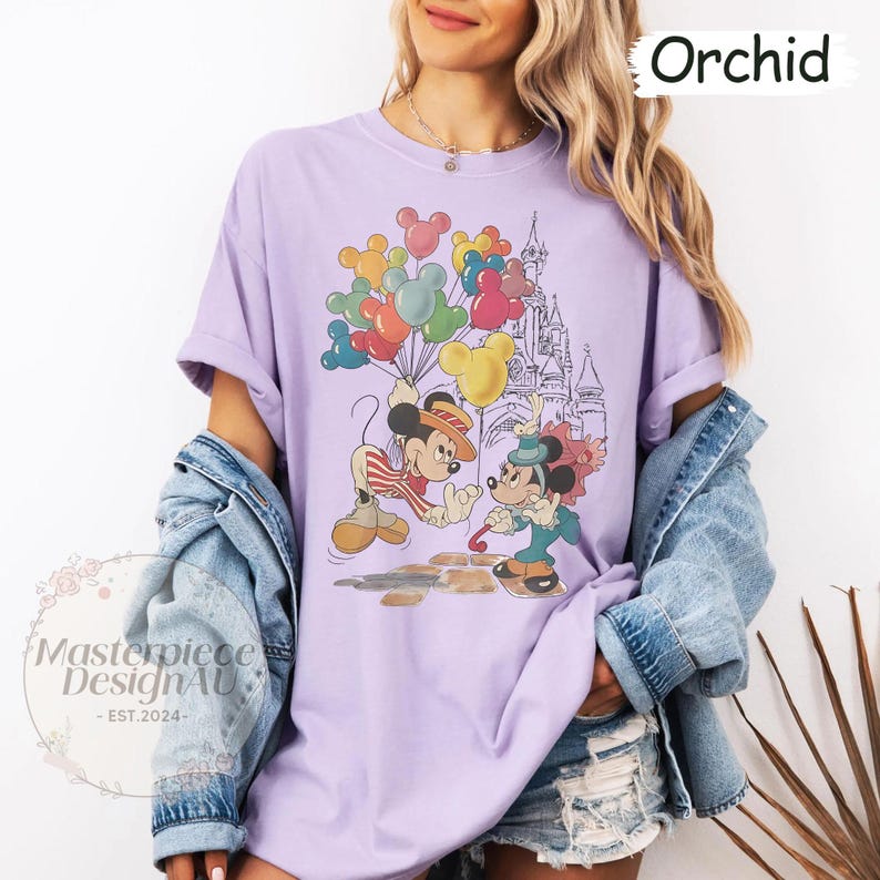 May include: A purple t-shirt featuring a vintage-style illustration of Mickey Mouse and Minnie Mouse holding balloons in front of a castle. The text "Orchid" is in the top right corner of the image.