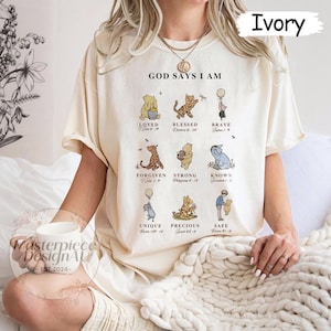 K&ouml;nnte beinhalten: Ein cremefarbenes T-Shirt mit dem Aufdruck "God Says I Am" auf der Vorderseite. Das Shirt zeigt Winnie Puuh-Charaktere mit Bibelversen, die unter jedem Charakter gedruckt sind. Zu den Charakteren geh&ouml;ren Winnie Puuh, Tigger, Ferkel, Esel und einige andere.