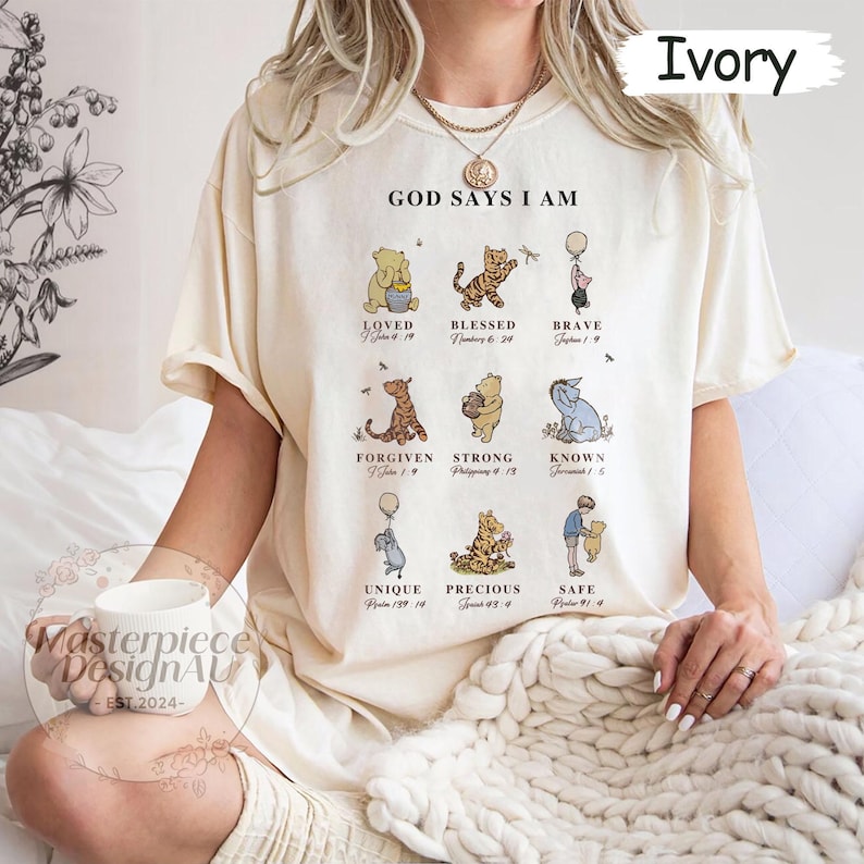 K&ouml;nnte beinhalten: Ein cremefarbenes T-Shirt mit dem Aufdruck "God Says I Am" auf der Vorderseite. Das Shirt zeigt Winnie Puuh-Charaktere mit Bibelversen, die unter jedem Charakter gedruckt sind. Zu den Charakteren geh&ouml;ren Winnie Puuh, Tigger, Ferkel, Esel und einige andere.