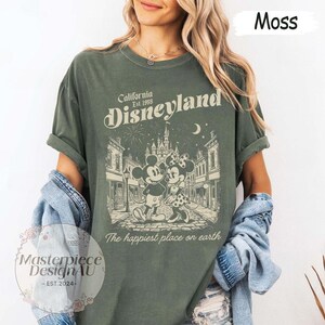 May include: A moss green short-sleeve t-shirt featuring a graphic print of Mickey and Minnie Mouse in front of a castle. The text reads California Est. 1955 Disneyland and The happiest place on earth. The design includes fireworks and a crescent moon.