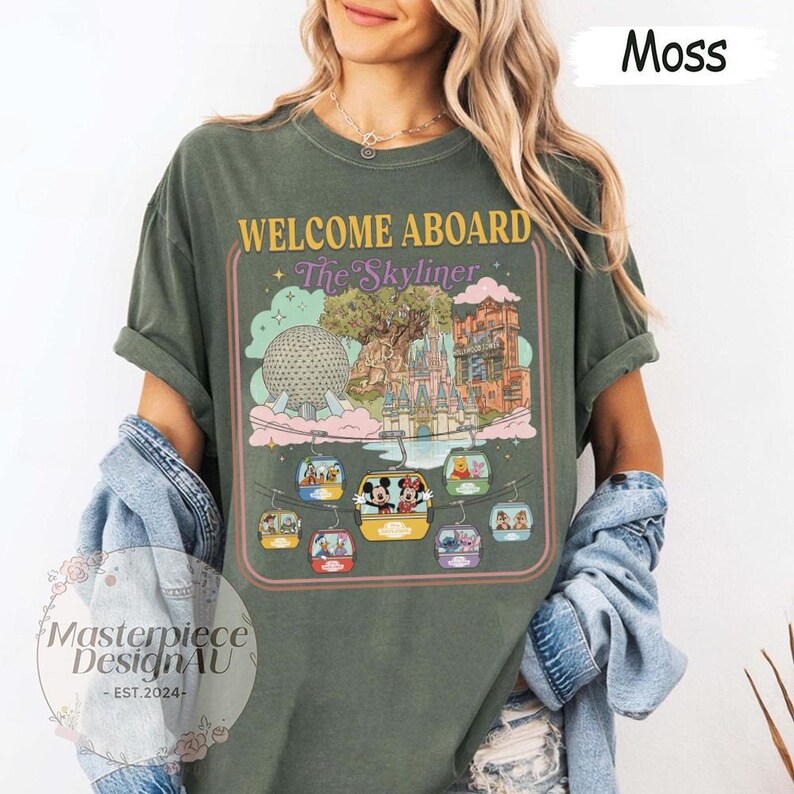 May include: Moss green t-shirt with a graphic featuring the text "WELCOME ABOARD The Skyliner" and illustrations of Disney theme park landmarks and characters. The design includes a retro-style frame and images of gondolas.