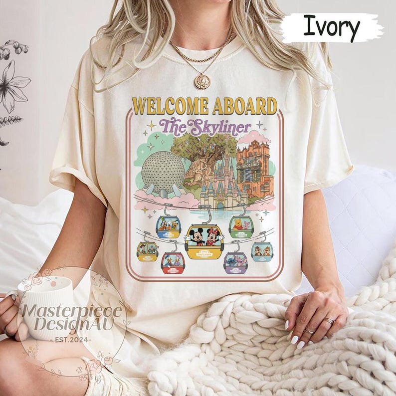 May include: Ivory-colored t-shirt featuring a colorful graphic of a theme park with the text "WELCOME ABOARD The Skyliner." The graphic includes images of the park's attractions and characters. The shirt is a relaxed fit.
