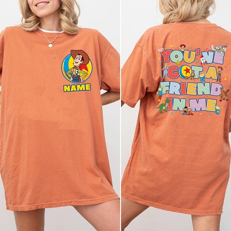 May include: An orange short-sleeve t-shirt featuring a front graphic of Woody inside a circular frame with the text NAME below. The back displays various Toy Story characters and the colourful text YOU'VE GOT A FRIEND IN ME.