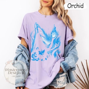 May include: Lilac t-shirt with a blue illustration of mythical figures and flames. The shirt has rolled-up sleeves and is worn with a denim jacket. The word "Orchid" is displayed in the top right corner.